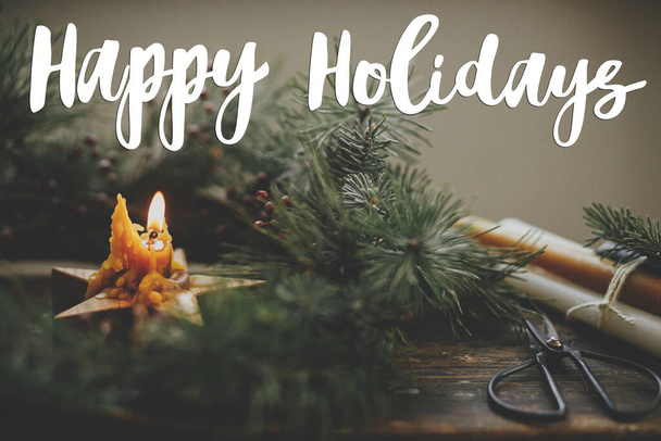 Happy Holidays κείμενο για κομψό κερί αστέρι καύση σε στεφάνι Χριστουγέννων με κλαδιά ερυθρελάτης και μούρα σε ρουστίκ ξύλο. Η κάρτα χαιρετισμού της σεζόν. Χειρόγραφο σημάδι. Καλά Χριστούγεννα.! - Φωτογραφία, εικόνα
