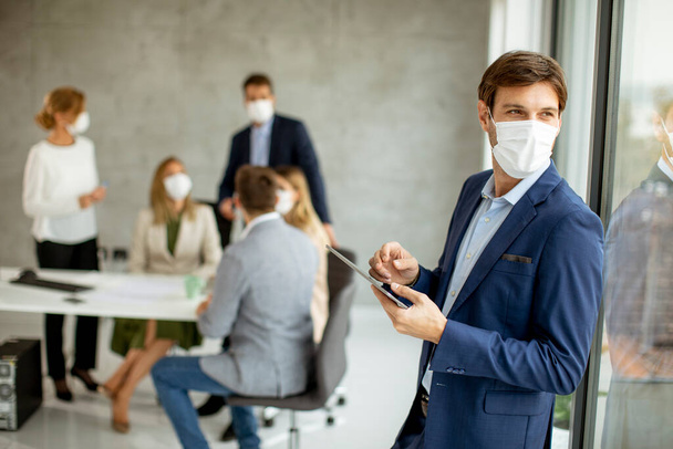 Bonito jovem homem de negócios usando máscara facial protetora enquanto segurava tablet digital no espaço do escritório - Foto, Imagem