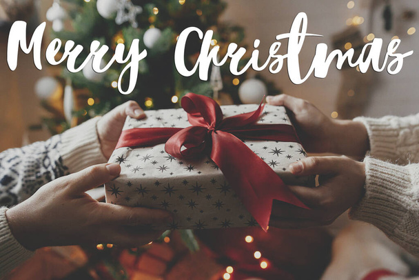 メリークリスマスグリーティングカード。クリスマスツリーの赤い弓とクリスマスプレゼントをライトと交換する手の上のメリークリスマステキスト。季節ごあいさつ。ハッピー・ホリデー！手書きのサイン - 写真・画像