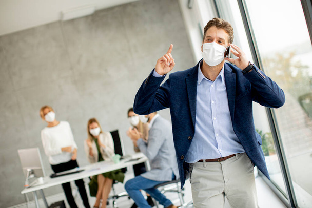 Schöner junger Mann benutzt Handy, während er Schutzmasken trägt, um Coronaviruus im Büro zu verhindern - Foto, Bild