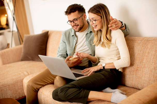 Bonito jovem casal usando laptop juntos enquanto sentado no sofá em casa - Foto, Imagem