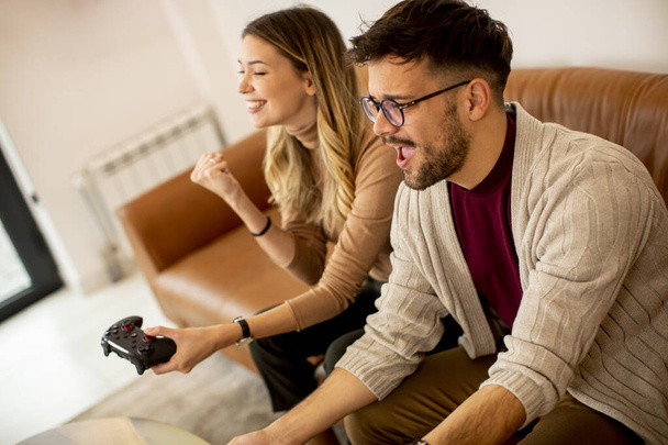Красивая молодая пара, играющая в видеоигры дома, сидящая на диване и развлекающаяся - Фото, изображение
