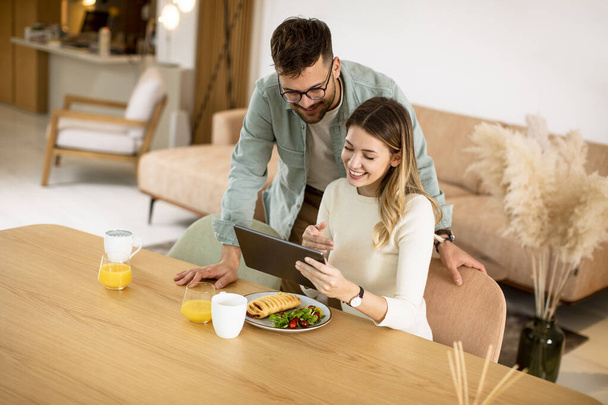 Lovely young couple using digital tablet and having breakfast in the kitchen - Фото, изображение