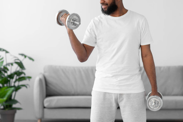 Mladý africký americký vousatý muž v bílém sportovním oblečení zvedá činky v obývacím pokoji interiéru, ořezané - Fotografie, Obrázek