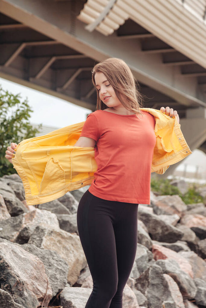 Pretty young sexy woman of athletic figure in bright clothes walking on coastline in city. Walk in city sea beach. Portrait of cute pretty female in yellow jacket and black pants on sea coast - Foto, afbeelding