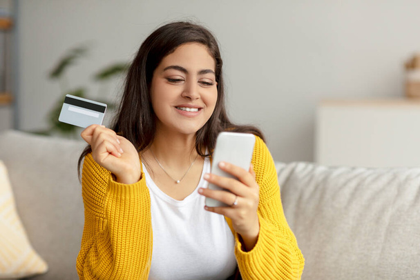 Online betaalconcept. Vrolijke arabische dame met creditcard en smartphone, zittend op de bank, het maken van internet aankoop - Foto, afbeelding