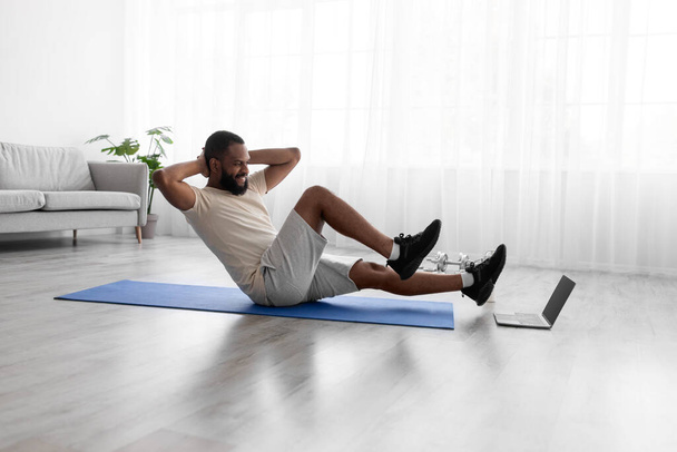 Feliz jovem afro-americano fazendo exercícios para pernas e abdominais no tapete no chão no interior do quarto - Foto, Imagem