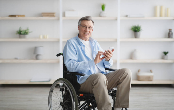 Hombre mayor positivo en silla de ruedas makin videollamada o navegar por la web en el teléfono inteligente en casa - Foto, imagen