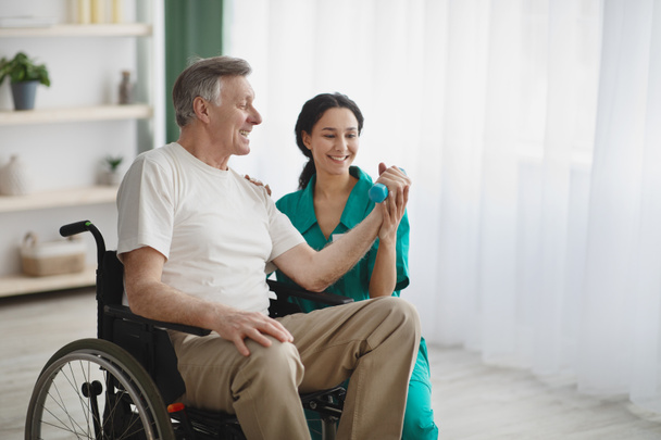 Jonge vrouwelijke fysiotherapeut helpt oudere man in een rolstoel om te trainen met halters, om te herstellen van letsel - Foto, afbeelding