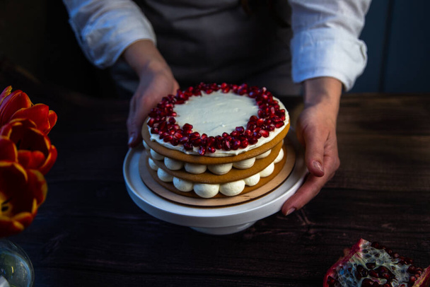 The cake stands on a white stand, decorated with cream and pomegranate on top, it is held by the hands of a pastry chef, there is a pomegranate next to it and there are flowers on a dark background - Photo, image