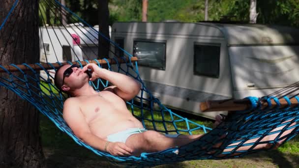 Een jongeman rust en praat aan de telefoon in een hangmat in de tuin.  - Video