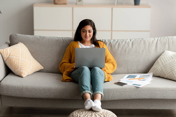 Jovem árabe freelancer feminino usando laptop usando fones de ouvido sem fio e digitando no teclado, sentado no sofá em casa - Foto, Imagem