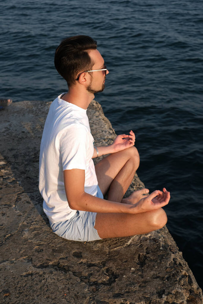 retrato de un joven en ropa deportiva medita en un muelle cerca del mar al amanecer. hombre deportivo se dedica a la meditación por el océano - Foto, Imagen