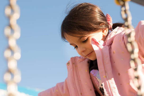 Маленький ребенок с длинными волосами, собранный в хвостик на фоне голубого неба в теплой одежде на детской площадке - Фото, изображение