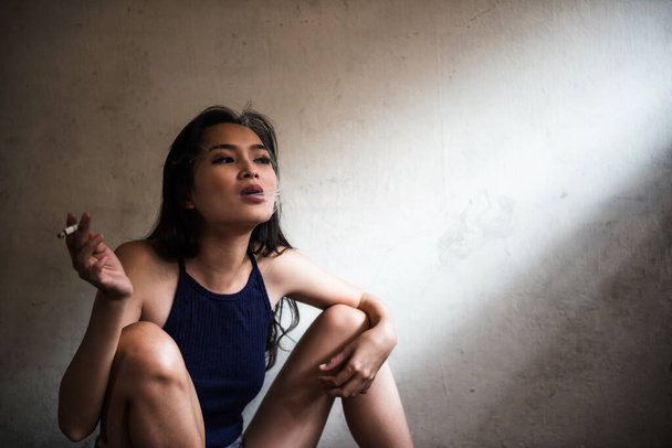 Азиатская молодая зависимая женщина курит сигарету возле стены грязной комнаты с солнечным светом через окно. Стрессовая девушка выдыхает дым. Здравоохранение и адаптация. - Фото, изображение