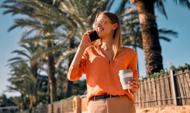 Eine junge Geschäftsfrau oder Studentin im Anzug trinkt Kaffee und telefoniert, während sie an der Küste vor der Palmenkulisse spaziert. - Foto, Bild