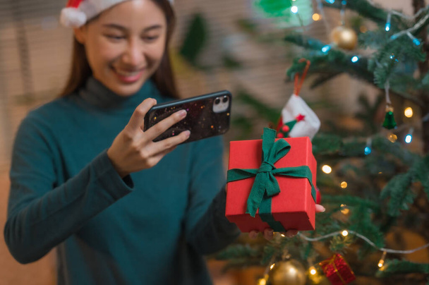 Felice donna asiatica utilizzando smartphone cellulare scattare foto di scatola regalo di Natale con albero di Natale in background - Foto, immagini