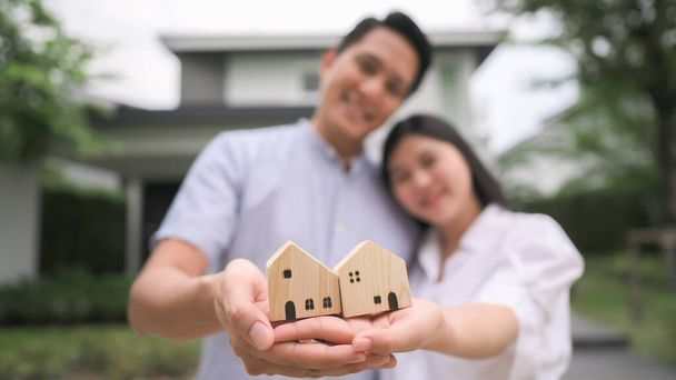Glückliches asiatisches Paar mit Holzhausmodell, das zusammen vor ihrem Haus steht. Neue Familie in neuem Haus gründen - Foto, Bild