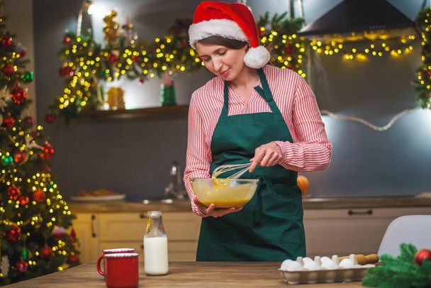A woman prepares food in the kitchen on Christmas Eve. A girl in an apron mixes the ingredients of the dough in a bowl . Merry Christmas and happy holidays. Decorated with Christmas lights interior. - Fotó, kép