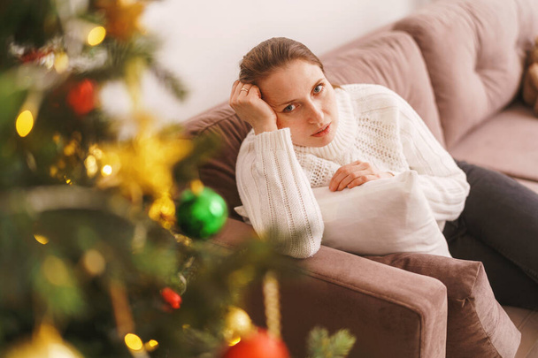 a frustrated young woman is sitting on the couch. the girl is upset, she has apathy and no desire. plans for the new year are collapsing. the concept of Christmas failures and loneliness - Foto, Bild