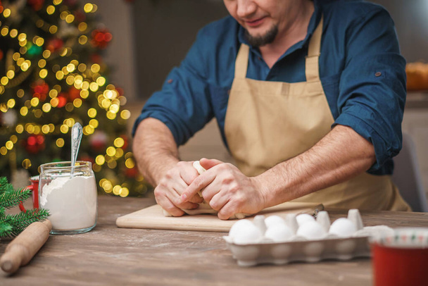 A man prepares food in the kitchen on Christmas Eve. A positive young adult man in an apron works with text and flour. Merry Christmas and happy holidays. decorated with Christmas lights interior. - Φωτογραφία, εικόνα