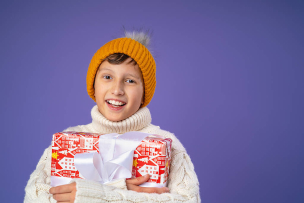 Happy laughing boy in knitting hat with Christmas gift on purple background. the child is happy and holds a big gift in his hands. New year sales, advertising, discounts. Copy space. - Foto, afbeelding