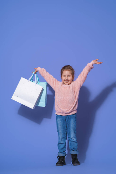 Stylowa dziewczynka w puszystych bluzach i dżinsach na fioletowym tle. papierowe torby z zakupami w rękach dziecka. Obraz emocjonalny. - Zdjęcie, obraz