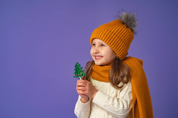 Little European girl of 5 years old with blonde hair in a yellow hat and scarf, holding a candy in the form of a green Christmas tree in her hand, stands smiling happily on a purple background. - Фото, зображення