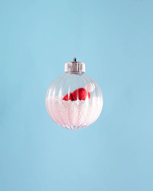 透明な装飾の泡、人工雪と内部のザンタクラウスの帽子。氷の青の背景に対する最小限の休日組成。メリークリスマスとハッピーニューイヤーグリーティングカード. - 写真・画像
