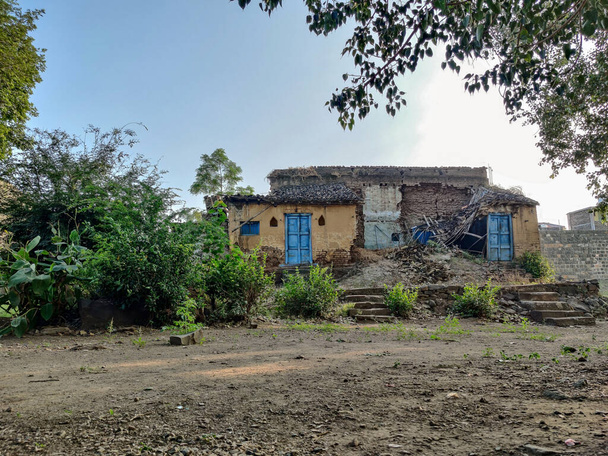 Фотографія пошкодженого, зруйнованого традиційного будинку червоної цегли з пірамідальним дахом у індійському селі, оточеного зеленими деревами. порожній простір перед домом у Хідрапурі (Махараштра, Індія).. - Фото, зображення