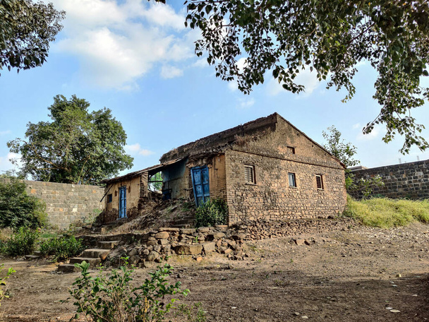 Фотографія пошкодженого, зруйнованого традиційного будинку червоної цегли з пірамідальним дахом у індійському селі, оточеного зеленими деревами. порожній простір перед домом у Хідрапурі (Махараштра, Індія).. - Фото, зображення
