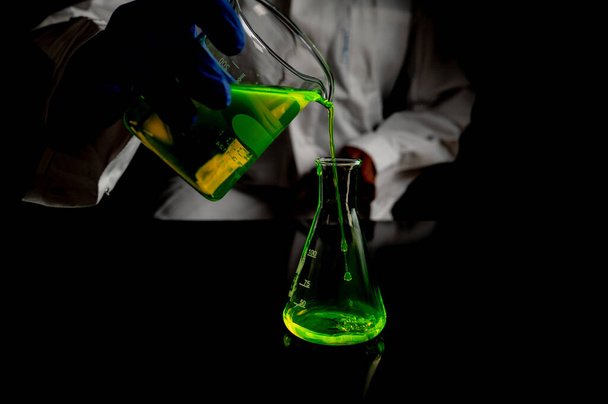 Ein Forscher experimentiert mit grünen fluoreszierenden Tröpfchen in einem Glaskolben in einem dunklen biomedizinischen Labor für die Entwicklung der Gesundheitsmedizin. Kopierraum schwarzer Hintergrund - Foto, Bild