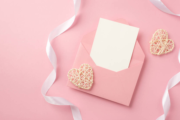Foto de vista superior de corazones de ratán sobre rosa abierto con hoja de papel y cinta de seda blanca sobre fondo rosa pastel aislado con espacio en blanco - Foto, Imagen