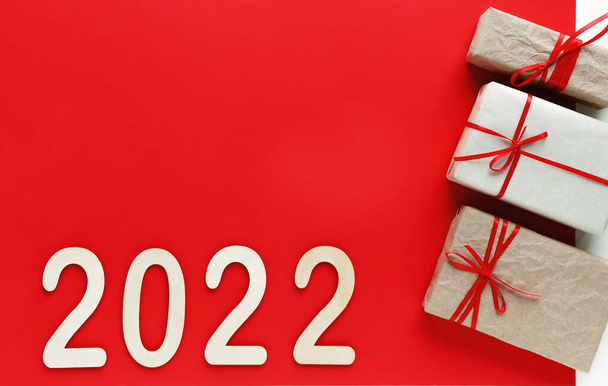 In a flat lay, top view, homemade gifts wrapped in craft paper and tied with a red ribbon are positioned on the right side and wooden numbers for the upcoming new year 2022 lie on a red background. - Foto, Imagem