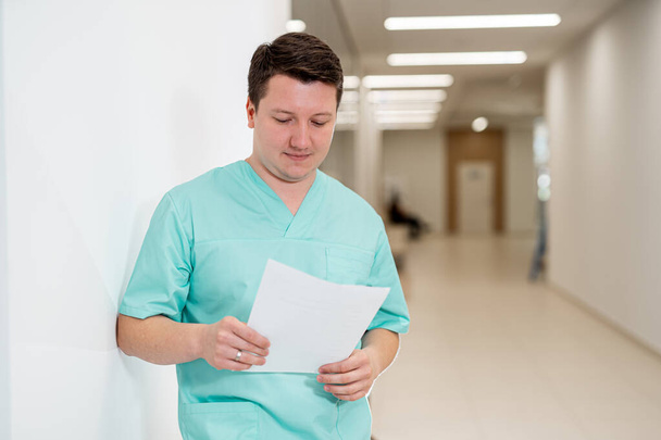 Doctor in blue clothes posing to the camera while reading documents. Medic standing near the surgery room. Vertical view - Foto, afbeelding