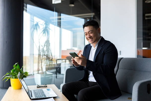 Successful Asian businessman celebrates victory, looks at phone, happy and rejoices sitting in modern office - Zdjęcie, obraz