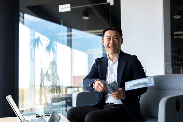 Happy and successful Asian, businessman working in office with laptop, happy holding cash dollars, throwing up, celebrating win and victory - Fotó, kép