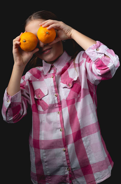 Piccola ragazza carina in posa con mandarini in studio. Foto isolata su sfondo nero con una sorgente luminosa. Il bambino sta scherzando chiudendo gli occhi con i mandarini.. - Foto, immagini