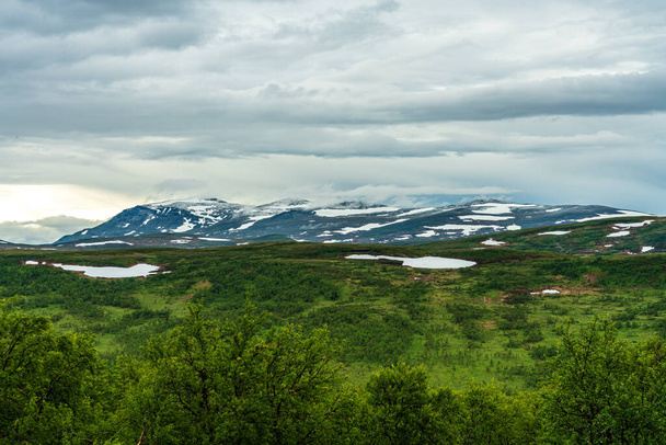 Hermosa vista de verano desde la inmensidad de las tierras altas suecas, vegetación verde de bajo crecimiento con manchas de nieve restantes y cumbres de montañas cubiertas de nieve en la distancia - Foto, Imagen