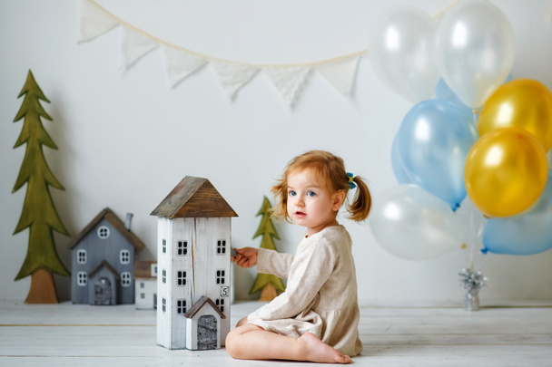Een klein meisje met twee paardenstaarten in een grijze gebreide jurk zit op de vloer en speelt met een houten huis. - Foto, afbeelding