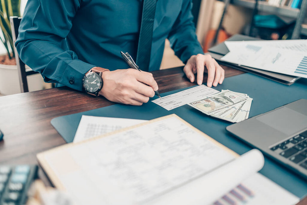 Business man hand writing and signing white blank bank check book and dollar bill, coin, laptop and graph chart on the desk at office. Payment by check, paycheck, payroll concept. - Valokuva, kuva