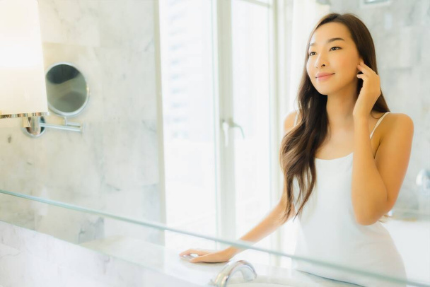 Retrato hermosa joven asiática mujer check up y maquillaje su cara en espejo en baño interior
 - Foto, Imagen