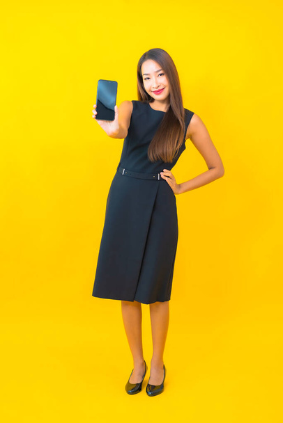 Portret mooie jonge Aziatische zakenvrouw met koffiebeker en slimme mobiele telefoon op gele achtergrond - Foto, afbeelding