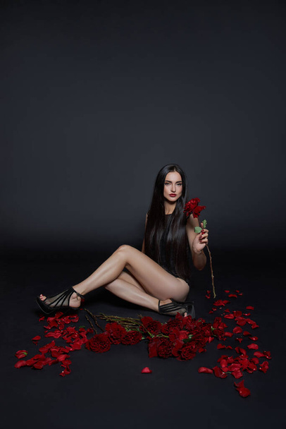 Beautiful brunette woman with a bouquet of rose flowers is sitting on the floor in a black bodysuit. Long hair, slim body - 写真・画像