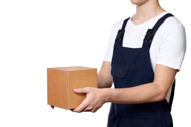 Body of man holding out a cardboard box in front of him and hands it over, isolated on white background. Man with parcel dressed in dark blue overalls and and a white T-shirt. Delivery concept. - Photo, image