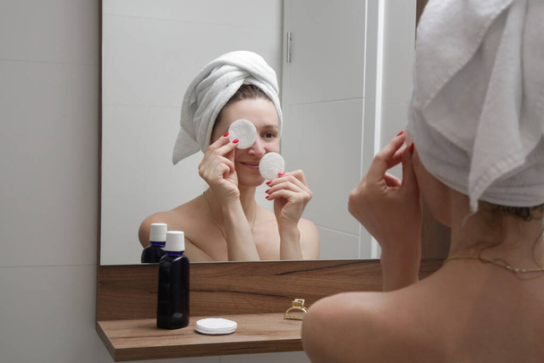 Retrato de uma bela jovem mulher de pé no banheiro e limpar o rosto com almofadas de algodão na frente do espelho, removendo maquiagem, beleza e conceito de spa. - Foto, Imagem