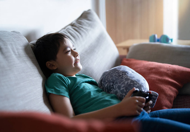 Portrait of happy kid holding a video game control. Child playing a online game at home,Young boy siting on a sofa having fun playing with a toy and relaxing on the weekend, New lifestyle after covid - Photo, Image
