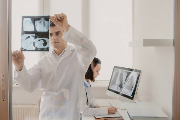 Lääketieteellisessä huoneessa kaksi nuorta lääkäriä radiologit analysoida röntgen- ja tietokonetomografia elokuva ja tehdä lääketieteellinen kuvaus kannettavan - Valokuva, kuva