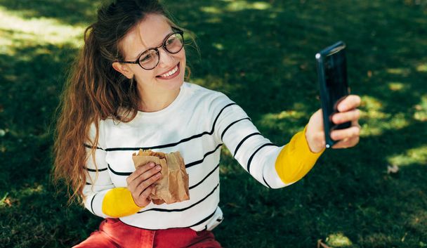 Smiling female taking selfies on the mobile phone during lunch with healthy food sitting on the green grass at college campus. A gorgeous young woman has online conversations on the smartphone. - Foto, Imagen