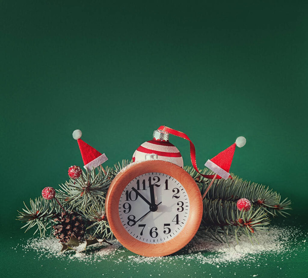 Countdown to midnight. Wood alarm clock with red baubles, Santa hats, berries, fir twigs and pine cones. Green paper background with snow. Copy-space, place for text. - Fotografie, Obrázek